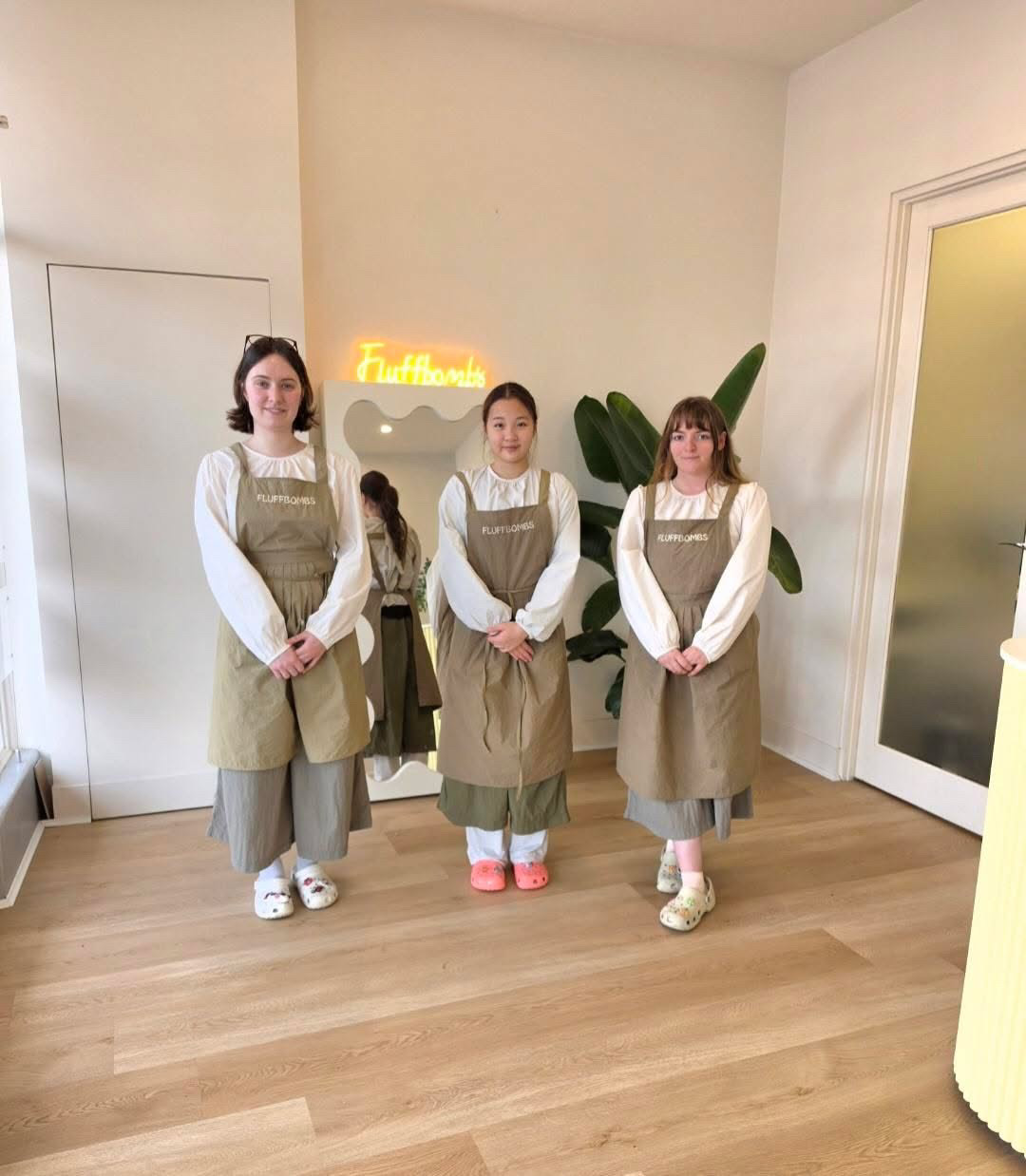 The Fluffbombs Grooming team in olive aprons in front of the salon's neon sign and wavy mirror